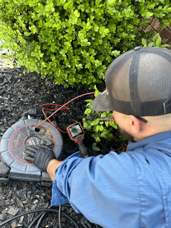 A man in a blue shirt and black hat looking at a small screen of a camera inside a sewage drain performing a plumbing pinpoint camera test.