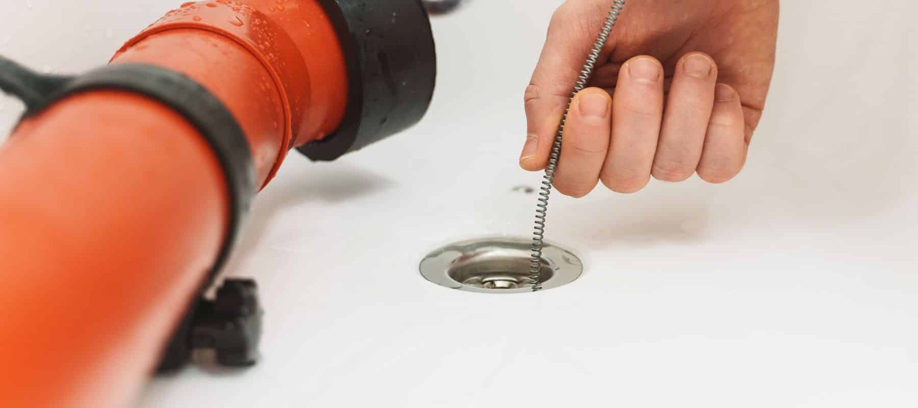 Close-up of a hand using a drain auger to clear a blockage, with a visible orange pipe and a clean sink drain