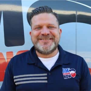 Jim's Plumbing Now team member wearing branded cap and company uniform, smiling in front of a company van
